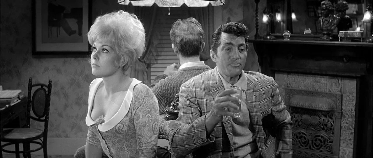 Kiss Me, Stupid (1964) movie still: medium — A 1960s living room features patterned wallpaper and a fireplace with decorative metal scrollwork. A…