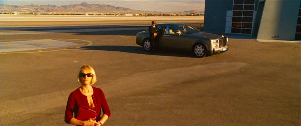 Ocean’s Thirteen (2007) movie still: wide — An open asphalt tarmac is flanked by desert mountains under a clear sky. A woman walks toward the camera…