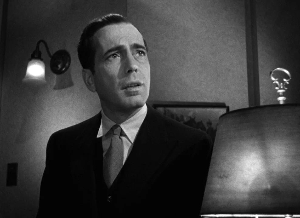 The Maltese Falcon (1941) movie still: medium — A dimly lit room with plain walls and a wall-mounted light fixture. A man in a dark suit gazes…
