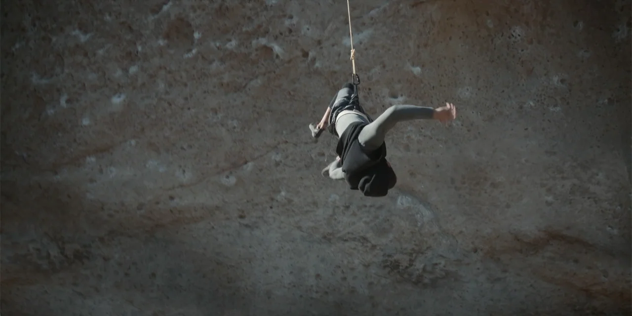 Fall (2022) movie still: wide — A high sandstone cliff face features deep textures and porous rock formations. A lone climber hangs on a rope in the…