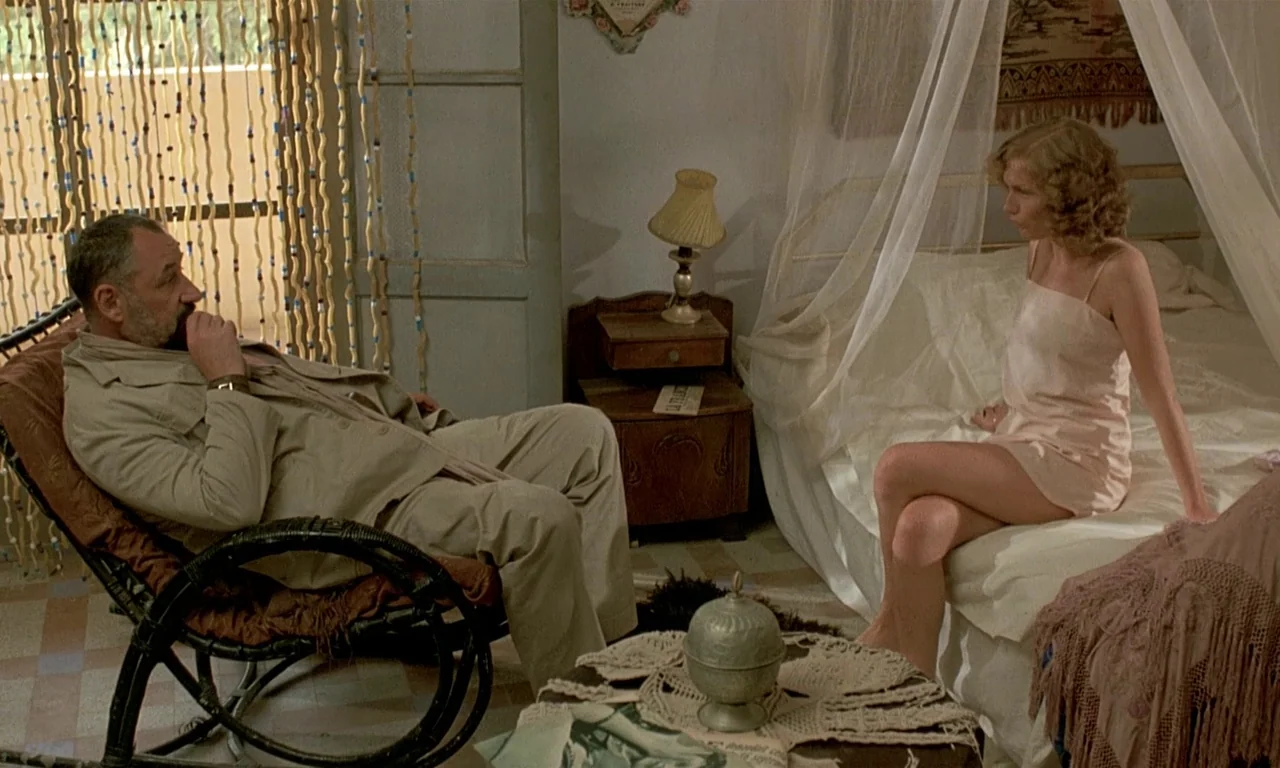 Coup De Torchon (1981) movie still: medium — A vintage sun-lit room features a four-poster bed and a bamboo rocking chair. A man sits in the chair…