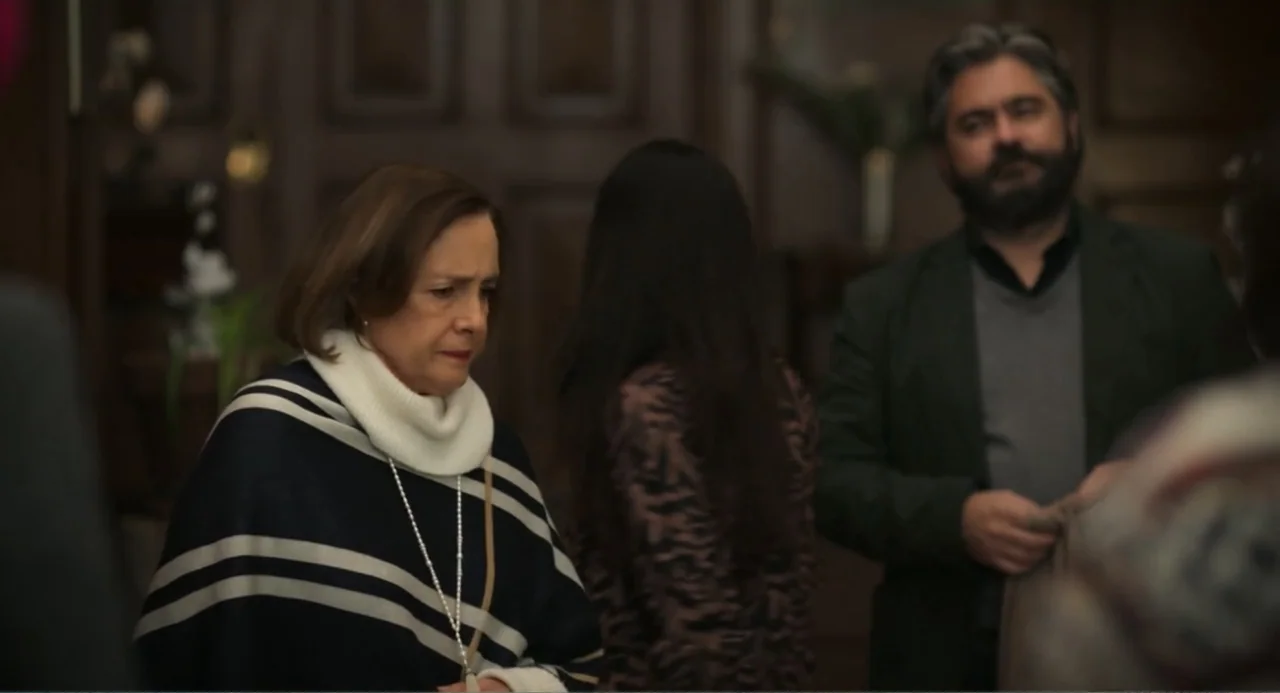La posada (2023) movie still: medium — An indoor room features wooden paneling and a dark, cluttered background with indoor plants. Three people…
