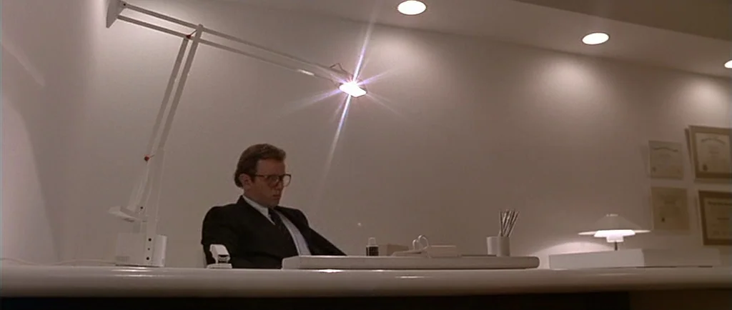 Manhunter (1986) movie still: medium — A minimalist, sterile private office features stark white walls and framed certificates. A man in a suit sits…