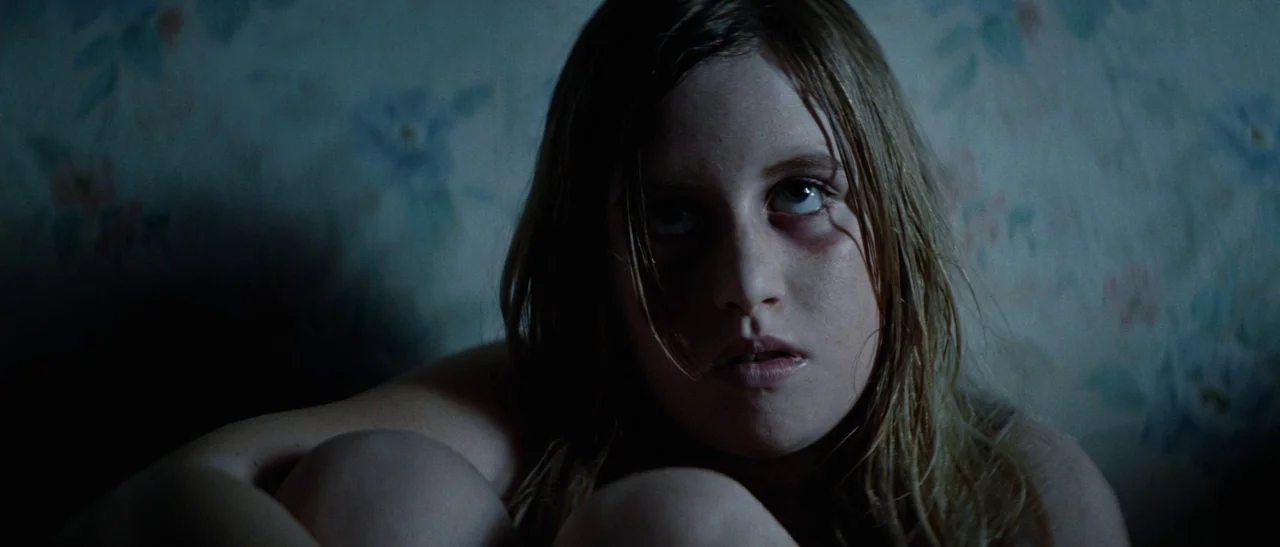 Outside Satan (2011) movie still: closeup — A dimly lit room features vintage floral patterned wallpaper showing signs of age. A young girl sits…