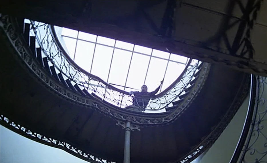 Possession (1981) movie still: wide — A grand spiral staircase features an ornate iron railing beneath a large skylight. A man stands on the upper…