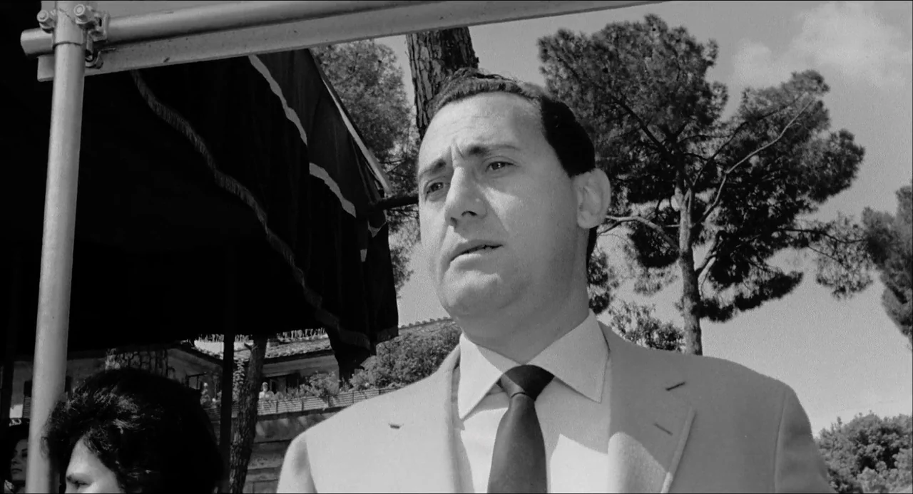 Il Boom (1963) movie still: medium — An outdoor park setting features large pine trees and a dark fabric awning canopy. A man stands in the…