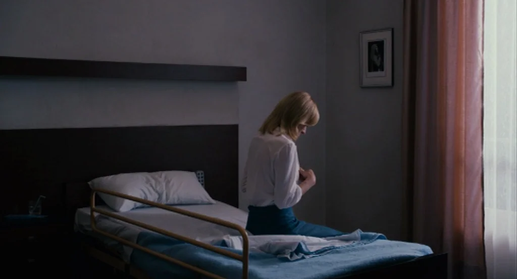 Lourdes movie still: medium — A spartan hospital room features a metal bed frame and a framed picture hanging. A woman sits on the edge of the bed…