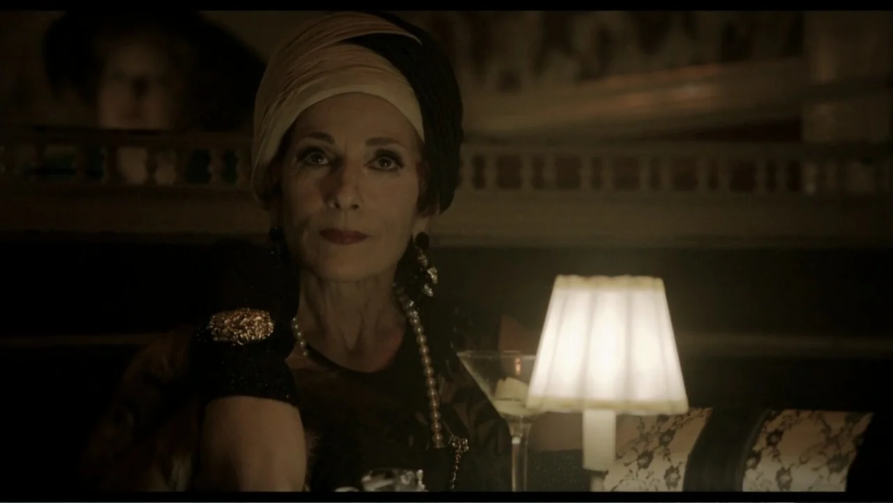 Six by Sondheim (2013) movie still: medium — A dimly lit interior setting features a woman seated at a table with a lamp. The woman wears a turban…