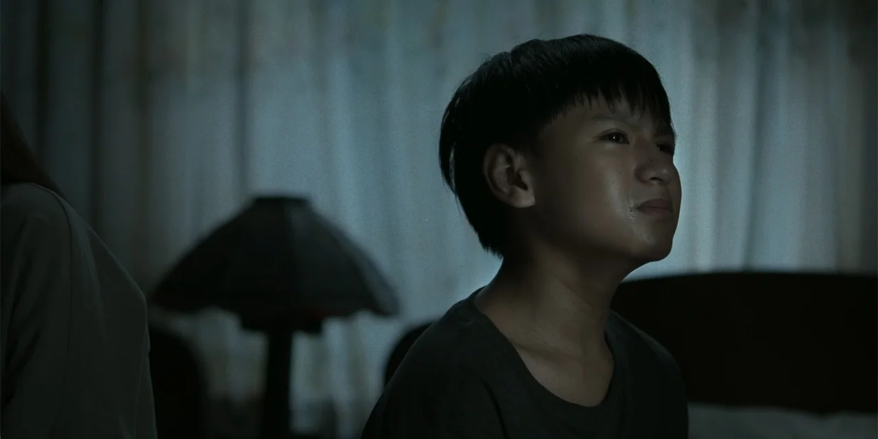 Nokturno (2024) movie still: closeup — An interior bedroom at night features dark walls and draped curtains in the background. A young boy in a dark…