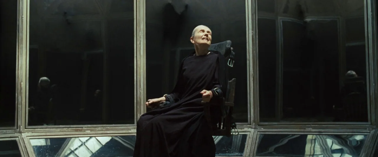 Mirrors (2008) movie still: medium — An angular, mirrored room with geometric panes reflecting the figure. A bald elderly woman sits in a wooden…