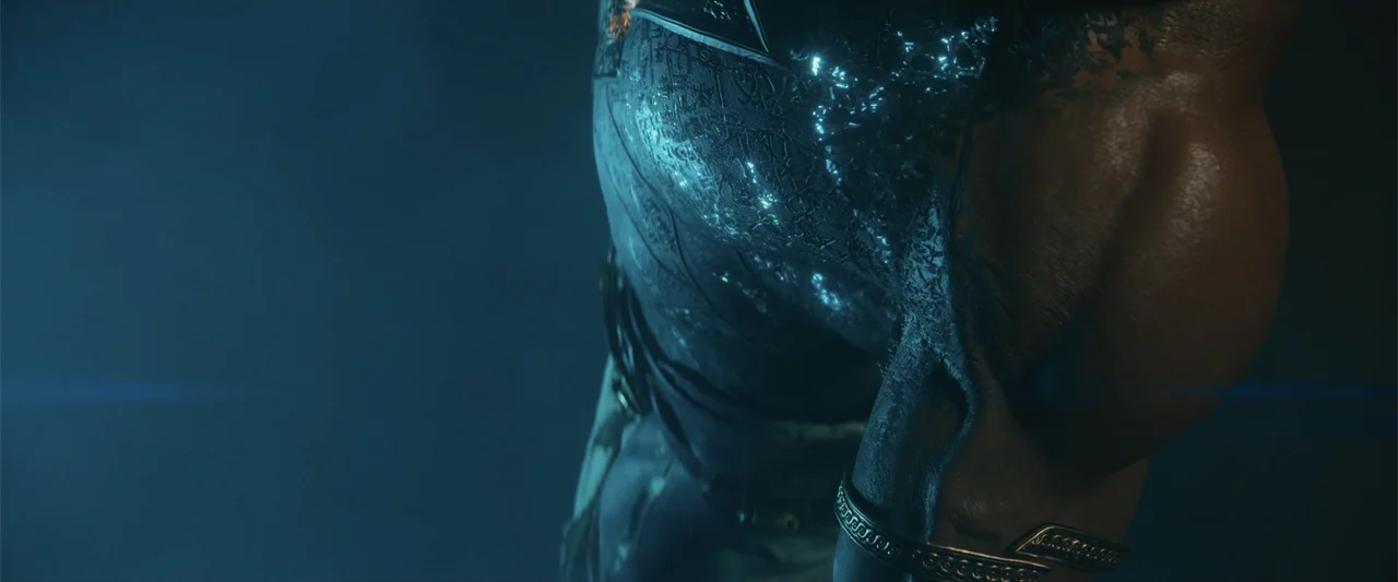 Black Adam (2022) movie still: closeup — A dark, undefined interior space focuses on the muscular shoulder and torso of a figure. The figure wears…