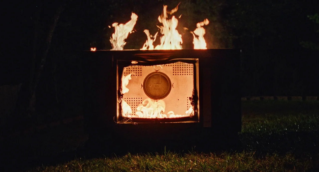 I Saw The TV Glow (2024) movie still: medium — An old cathode ray tube television sits on a dark grassy lawn at night. Bright orange flames emerge…