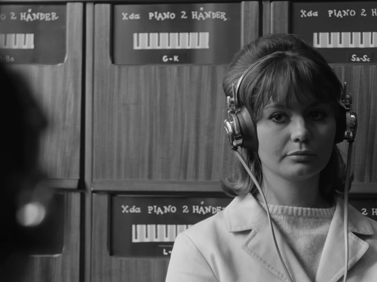 The Baby Carriage (1963) movie still: medium — A woman stands before several wooden filing cabinets labeled with music category signs. She wears…
