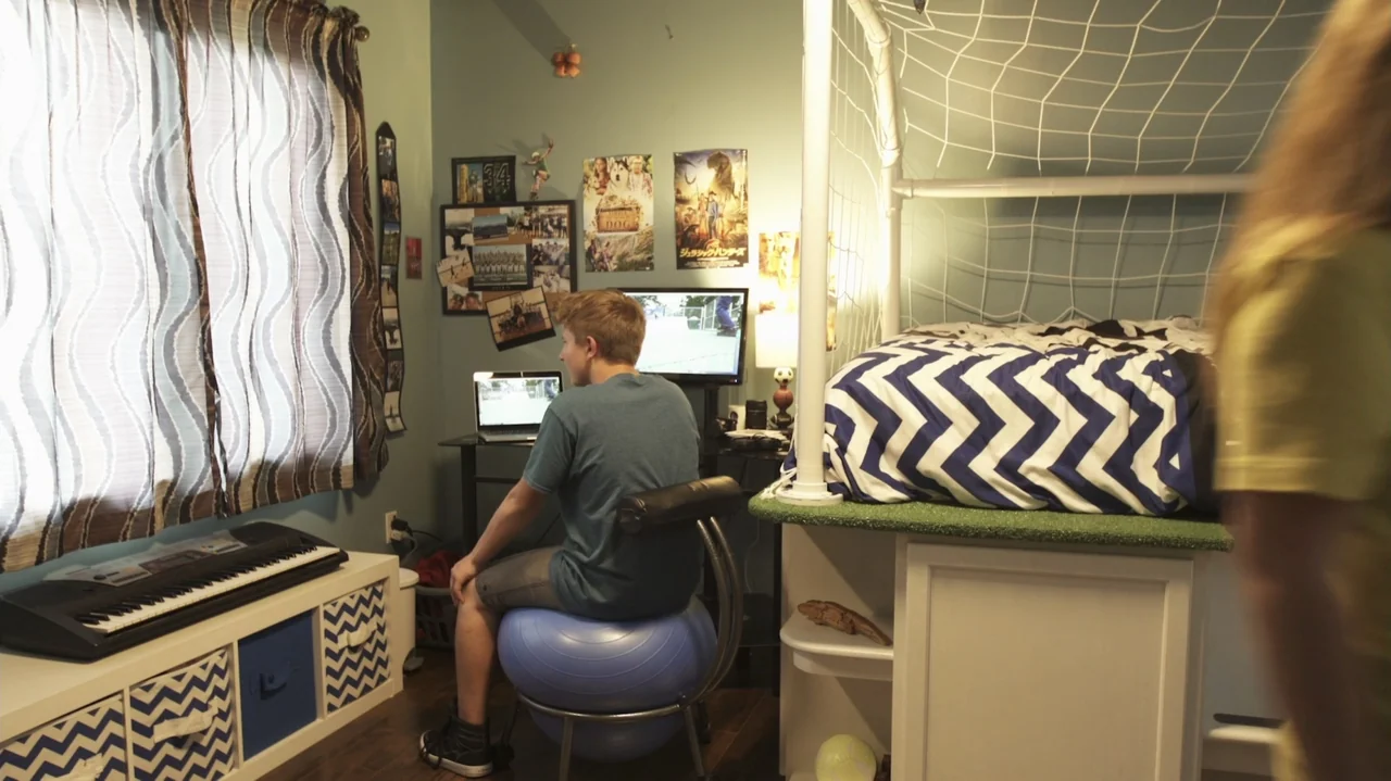 Sk8 Dawg (2018) movie still: medium — A cluttered teenager's room features a loft bed with a sports net structure. A boy sits on a yoga ball facing…