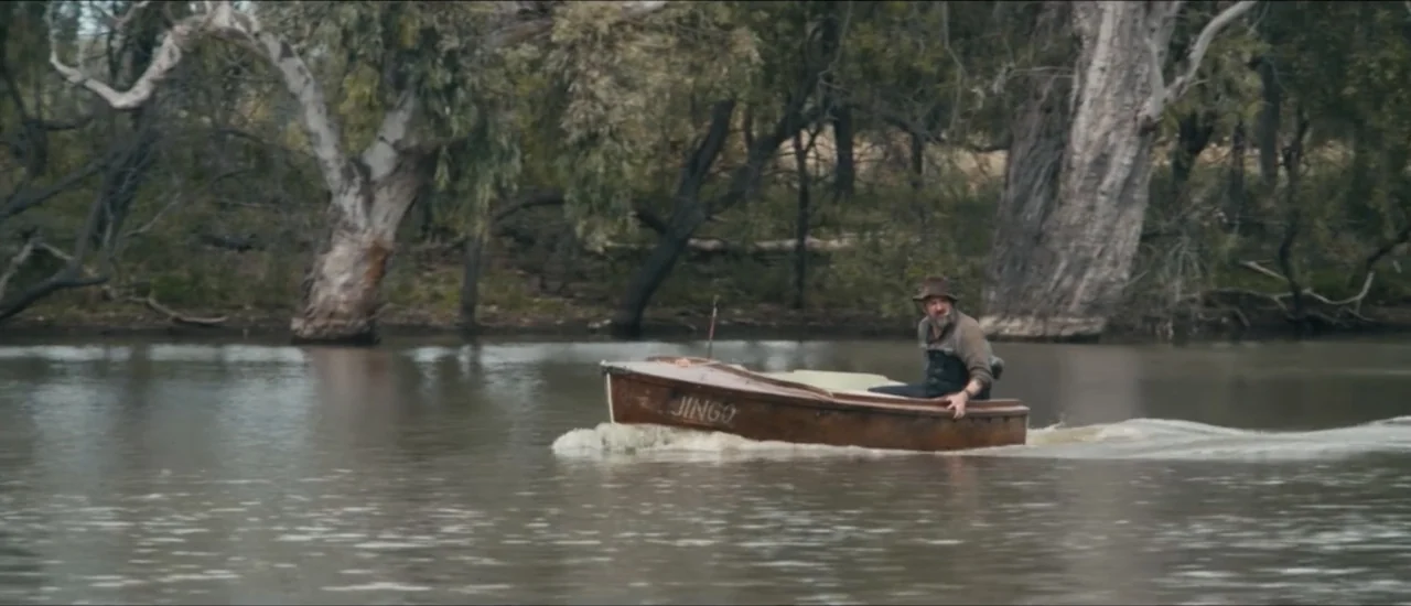 Fear Below (2025) movie still: wide — A murky river winds through a dense bank of large, grey-barked eucalyptus trees. A man sits in a small wooden…