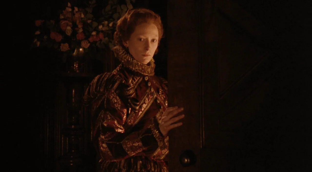 Orlando (1992) movie still: medium — A dark manor hallway features a tall flower arrangement on a pedestal in shadow. A woman dressed in historical…