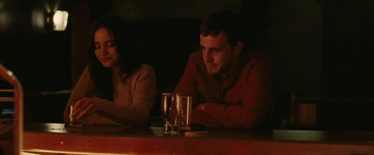 God’s Creatures (2022) movie still: medium — A dimly lit bar interior features a wooden counter and dark background shadows. A young couple sits…