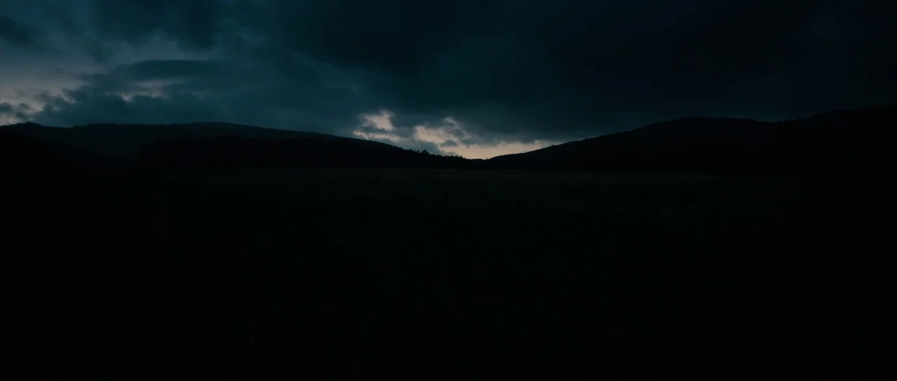 The Duke of Burgundy (2014) movie still: establishing — A vast, dark field stretches out beneath a heavy, cloud-filled night sky. Low rolling hills…
