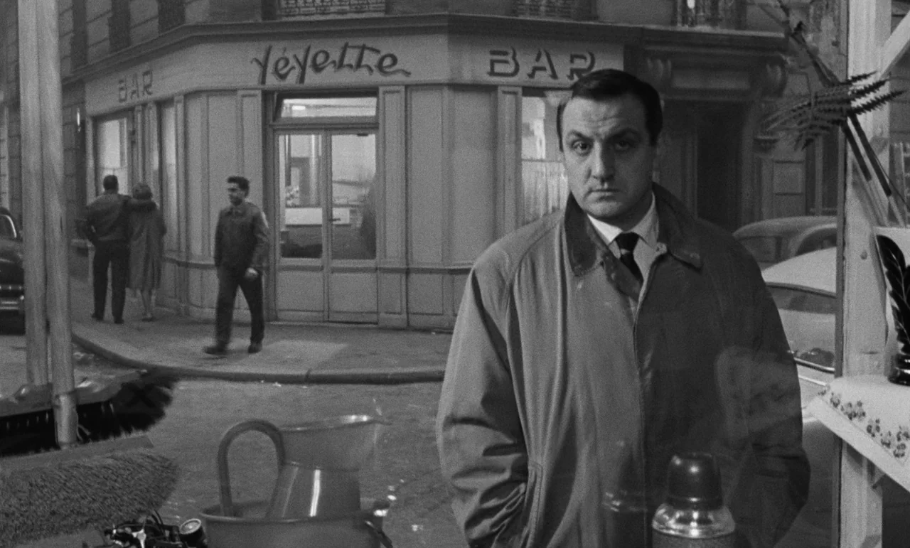 Witness In The City (1959) movie still: medium — A street corner in Paris features a cafe with the name Yeyette Bar painted. A man in a light…