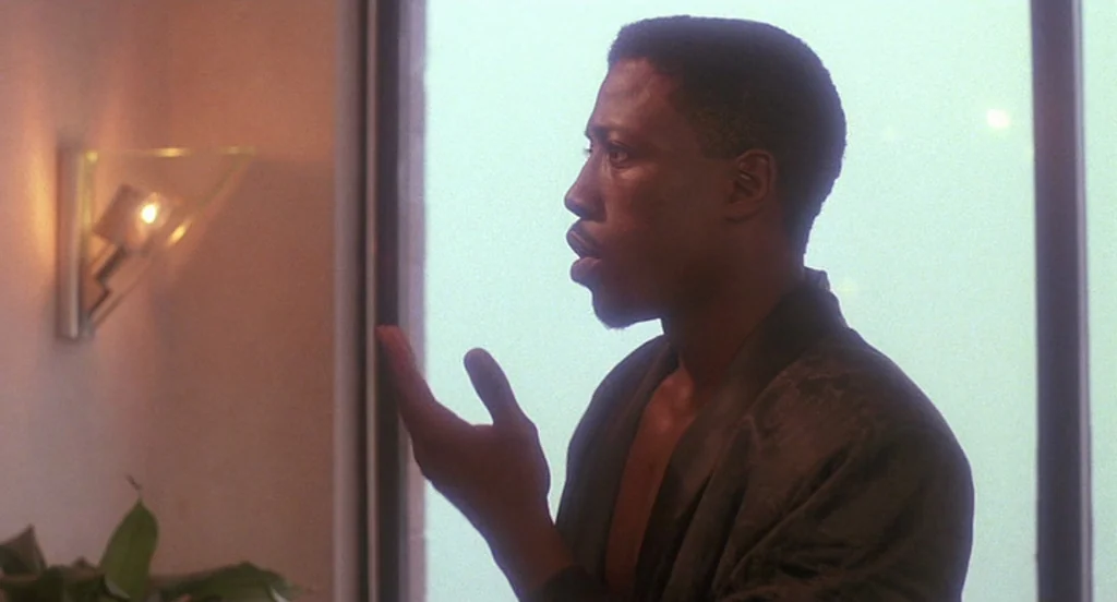 Mo’ Better Blues (1990) movie still: medium — A dimly lit room with a wall sconce and a large window behind the subject. A man in a robe gestures…