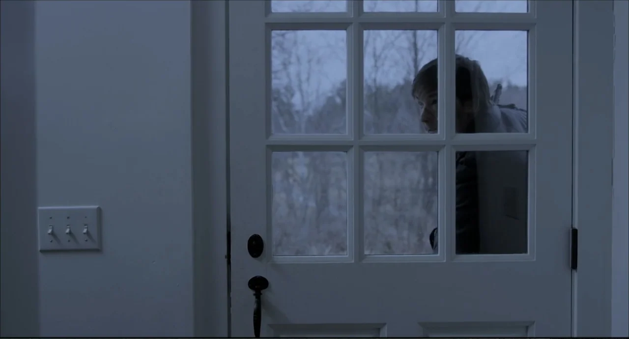Freedom (2024) movie still: medium — A white interior door with multiple glass panes separates an interior space from outside. A man stands on the…