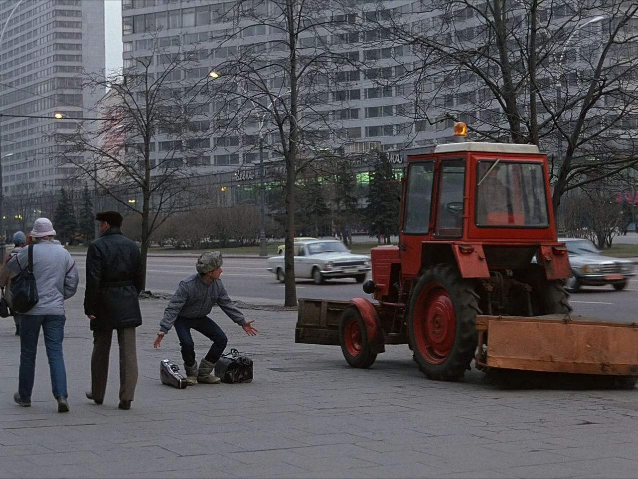 Kin-dza-dza! (1986) movie still: wide — A wide city street with a large red industrial tractor parked on the sidewalk. A man in a fur hat kneels…