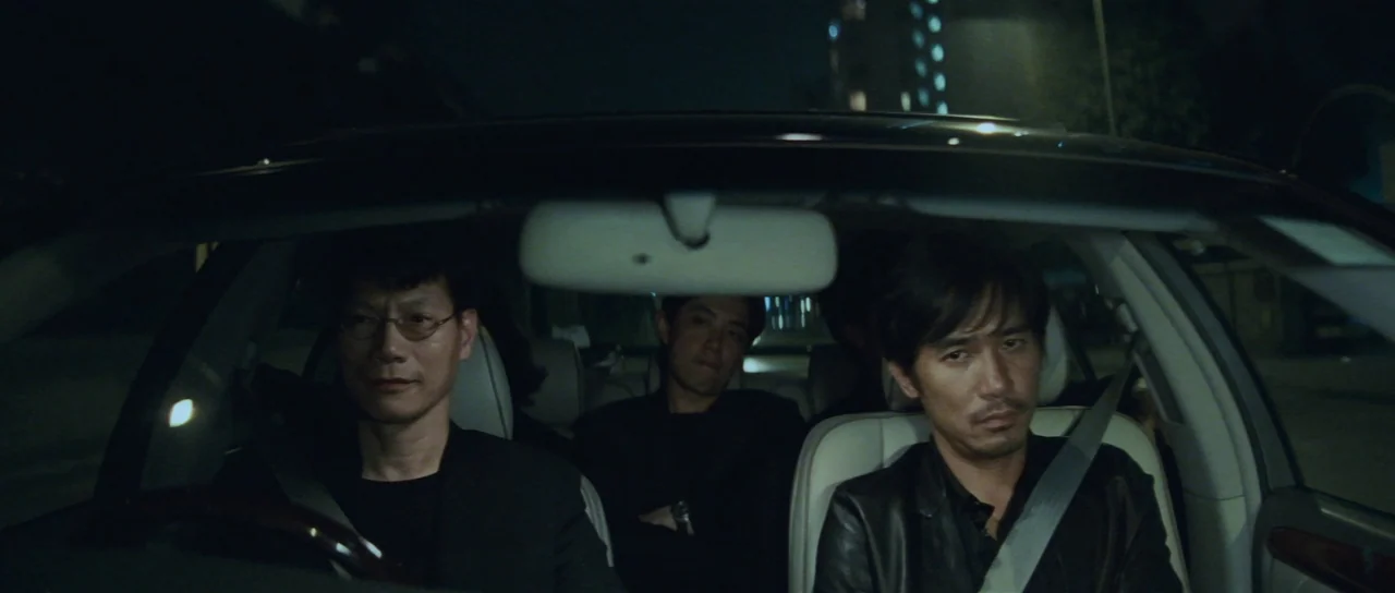 Infernal Affairs (2002) movie still: medium — The interior of a sedan at night carries three men in dark clothing. The driver operates the vehicle…