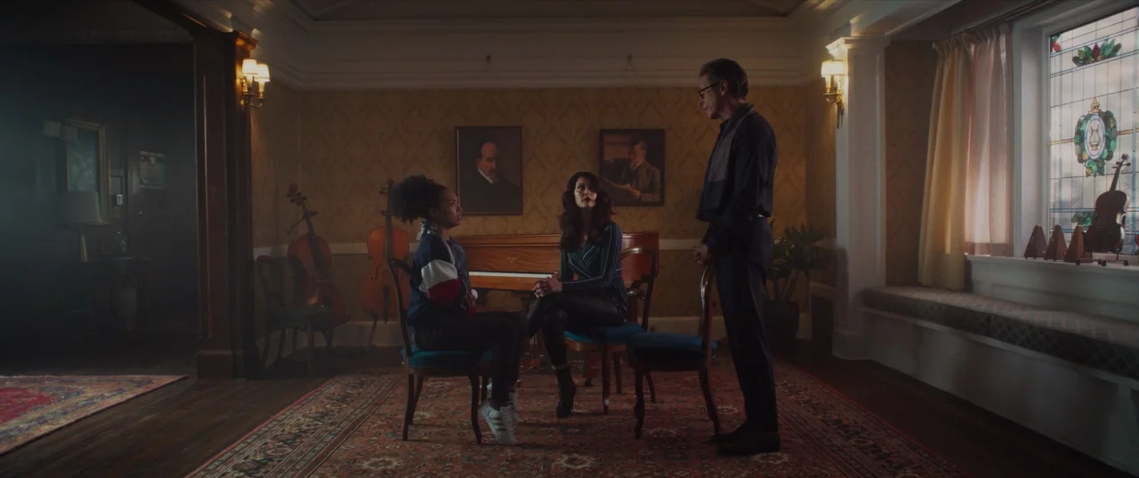 The Perfection (2018) movie still: wide — A stately music room features paneled walls, patterned rugs, and two large cellos. Two women and a man…