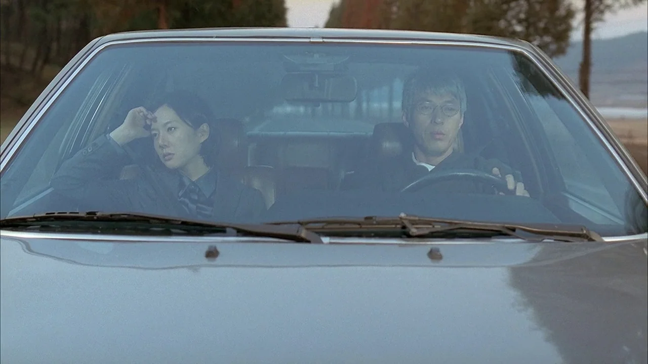 A Tale of Two Sisters (2003) movie still: medium — A man and a woman sit inside a car on a road lined with trees. The man holds the steering wheel…