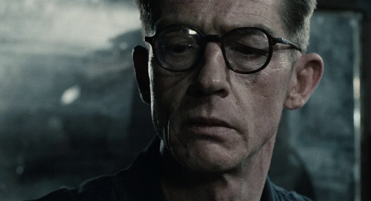 1984 (1984) movie still: closeup — A dimly lit room with muted grey tones and textured, out-of-focus background elements. A middle-aged man with…