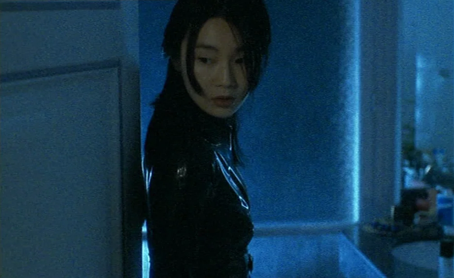 Irma Vep (1996) movie still: medium — A dark bathroom is illuminated by a glowing blue wall panel behind the subject. A young woman in a black…