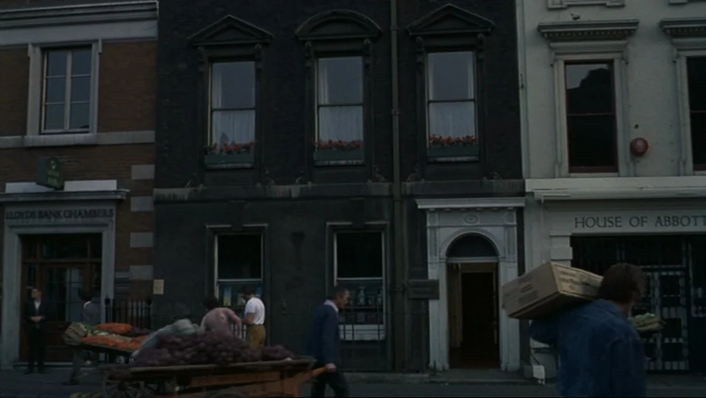 Frenzy (1972) movie still: wide — A row of London-style stone buildings lines a street with commercial signage at ground level. Pedestrians walk…