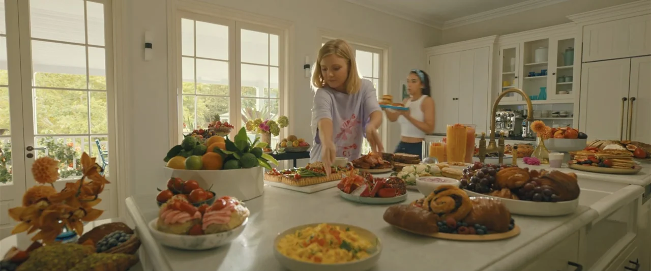 Anyone But You (2023) movie still: medium — A sunlit bright modern home with white cabinetry and a large marble island covered in food. A woman in a…