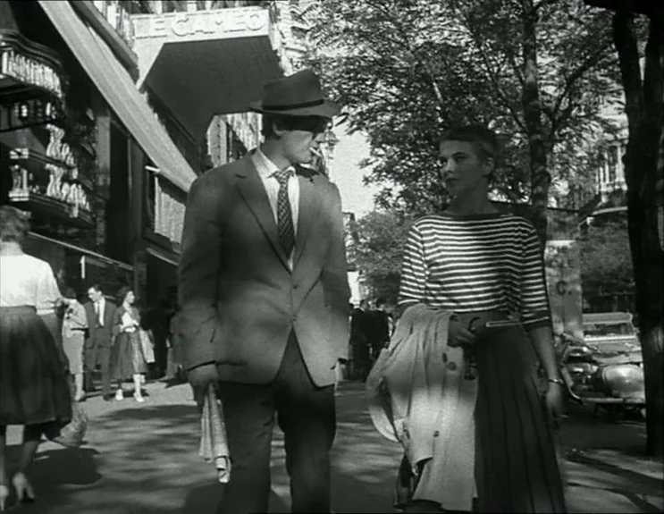 À Bout De Souffle (Breathless) (1960) movie still: fullbody — An urban sidewalk with trees and a cinema theater canopy in the background. A man…