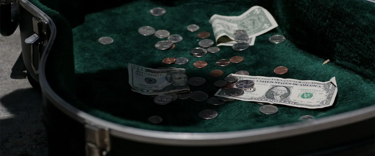 Playing God (2021) movie still: closeup — The open interior of a black guitar case is lined with plush green felt. Several US dollar bills and…
