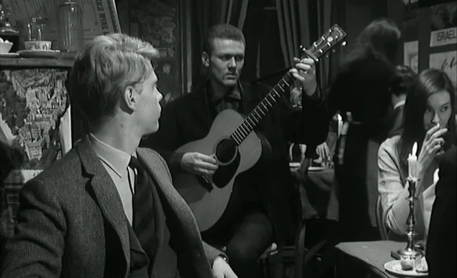 The Servant (1963) movie still: medium — A dimly lit European style establishment with dark walls and posters in the background. A man plays an…
