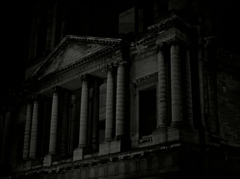Eloge de L’amour (2001) movie still: establishing — A dark, neoclassical building facade features multiple stone columns and a triangular pediment.…