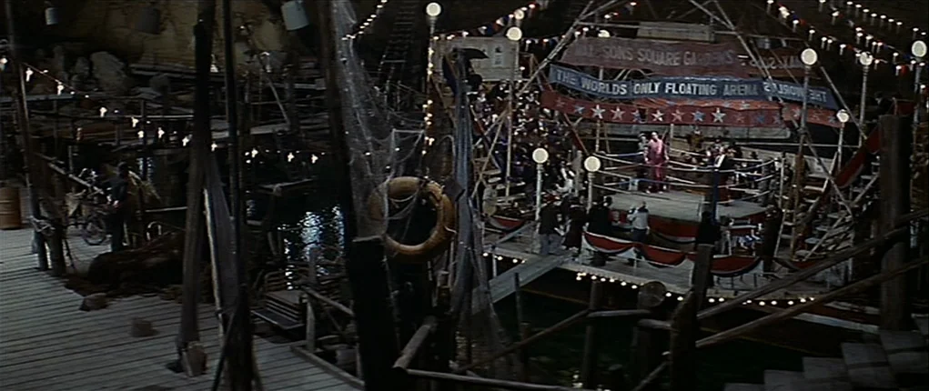 Popeye (1980) movie still: wide — A floating makeshift boxing arena built within a dark, water-filled grotto or cavern interior. A small group of…