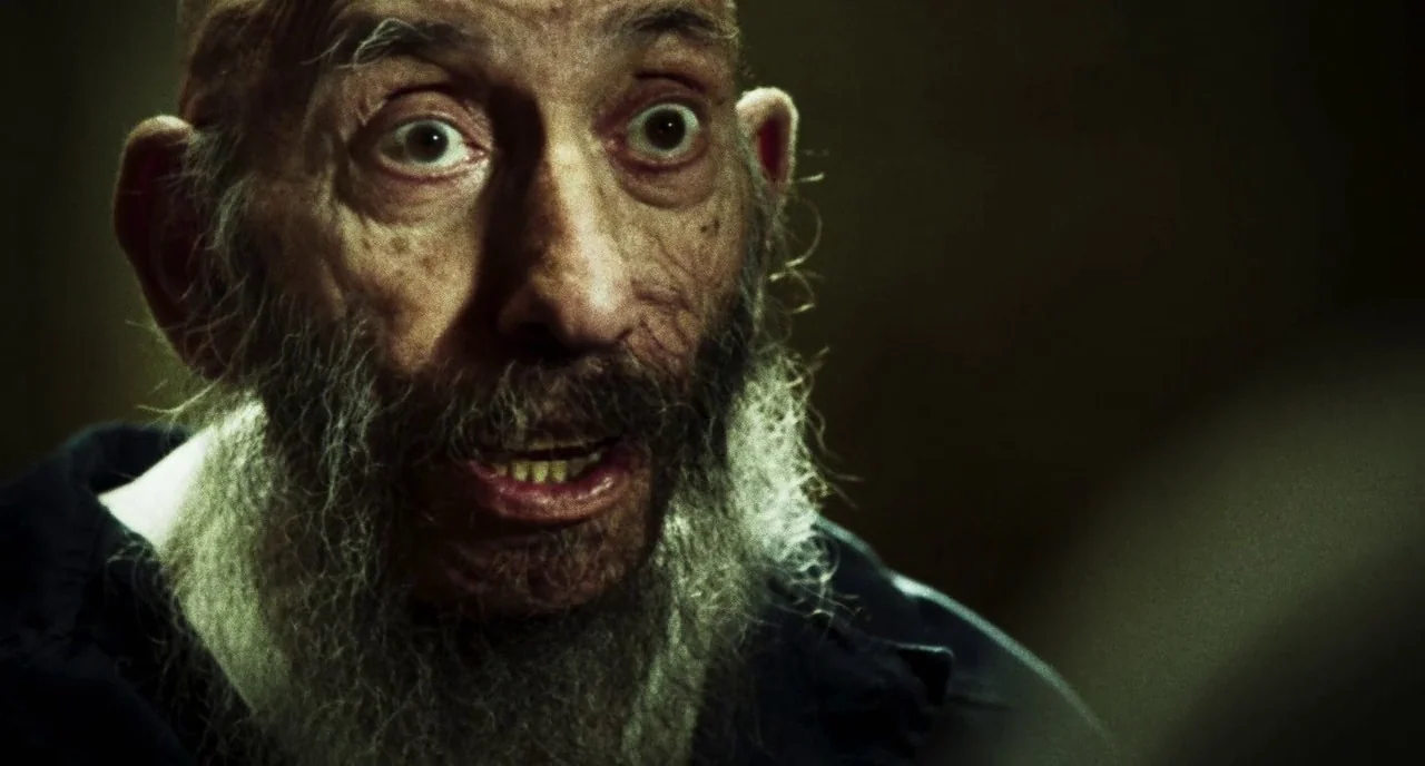 3 From Hell (2019) movie still: closeup — The face of an elderly man is centered in a dimly lit, dark space. The subject looks off-camera with wide,…