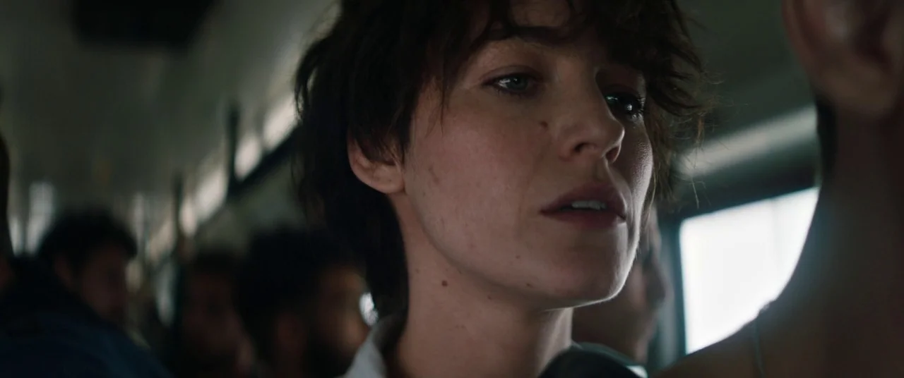 The Rhythm Section (2020) movie still: closeup — A crowded train car interior with blurred passengers seated in the background. A woman with short…