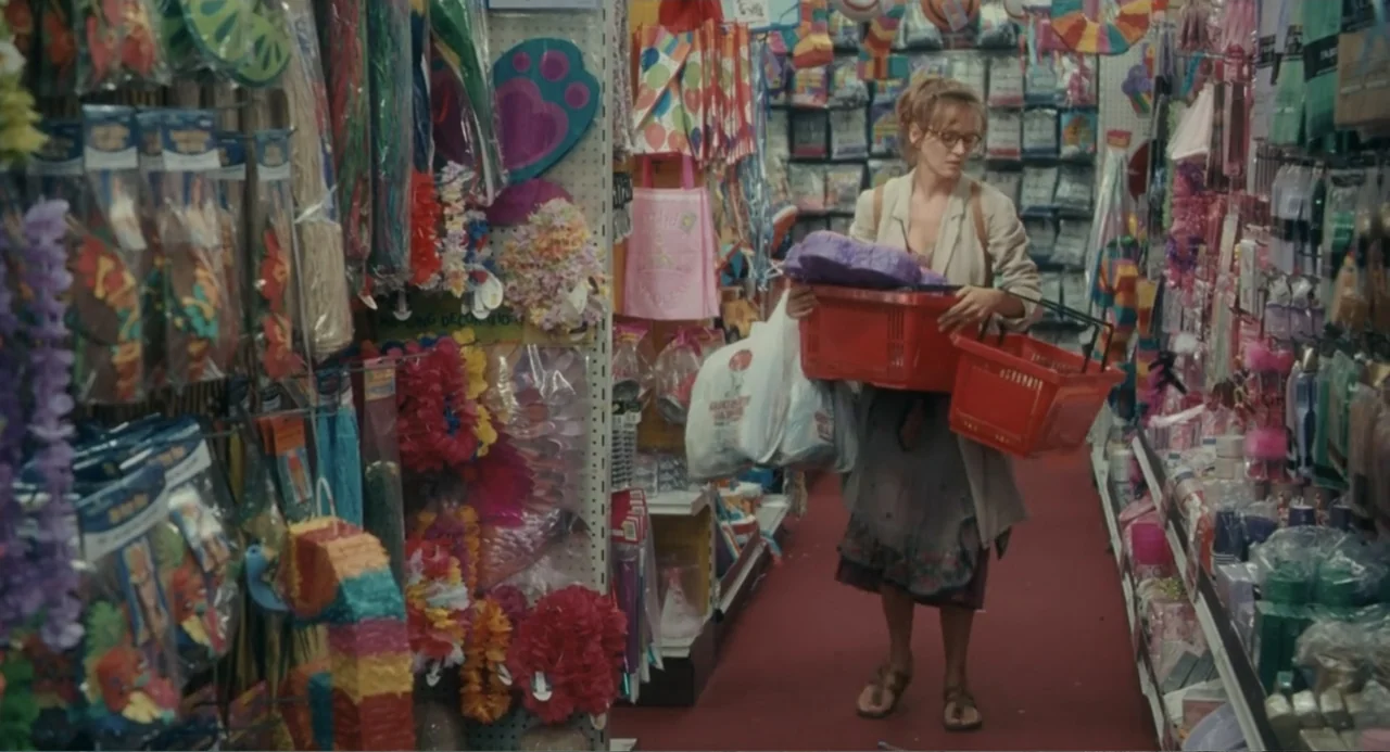 Motherhood (2020) movie still: fullbody — A cramped party supply store aisle is lined with overflowing shelves of colorful plastic party goods. A…