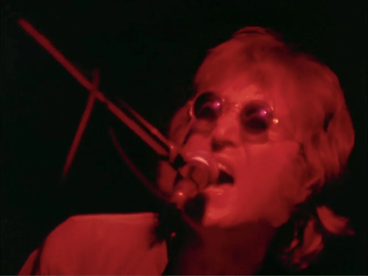 One to One: John & Yoko (2025) movie still: closeup — A dark concert venue with minimal visual information beyond the primary subject. A man sings…