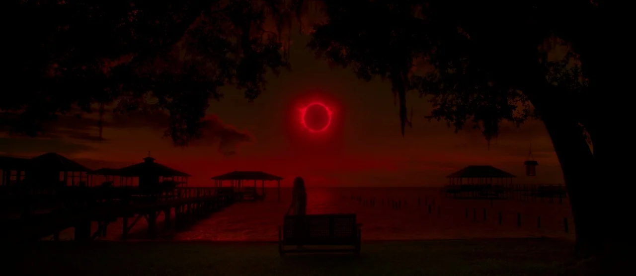 Gerald’s Game (2017) movie still: wide — A pier juts into dark water under a deep red eclipse sky. A woman stands alone near a wooden bench by the…