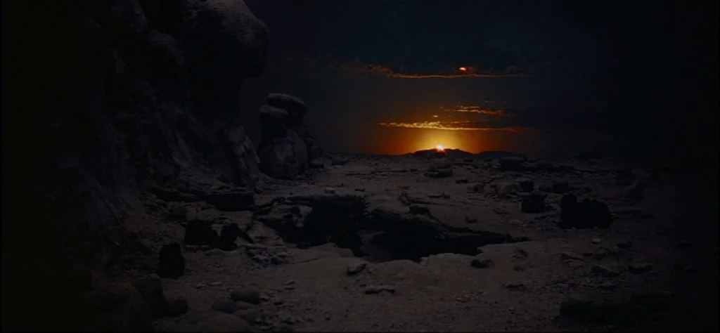 2001: A Space Odyssey (1968) movie still: establishing — A dark, rocky desert landscape stretches out under a dim, twilight sky. The sun is a bright…