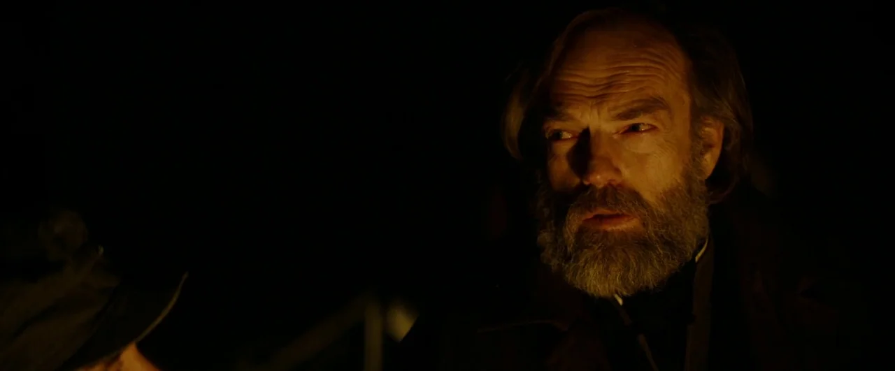 Black ’47 (2018) movie still: closeup — A dark outdoor setting at night illuminated by a nearby fire. A middle-aged man with a full beard looks…