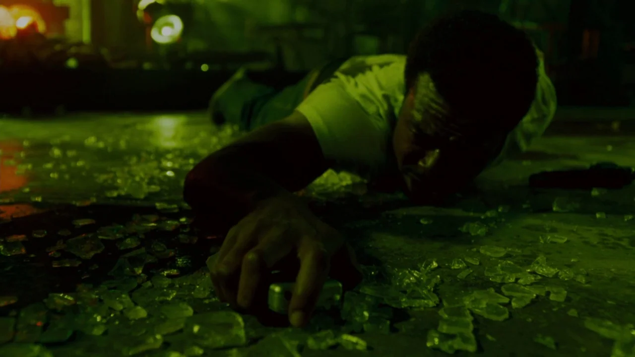 Saw IV movie still: medium — A dark industrial floor covered in fragments of broken glass and debris. A man lies prone on the ground reaching toward…