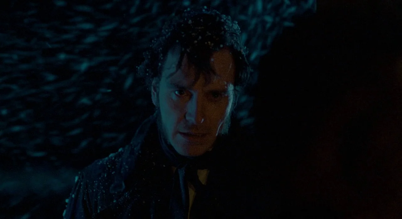 Jane Eyre (2011) movie still: closeup — A man stands in a dark, snow-filled night environment with minimal background detail visible. The subject…