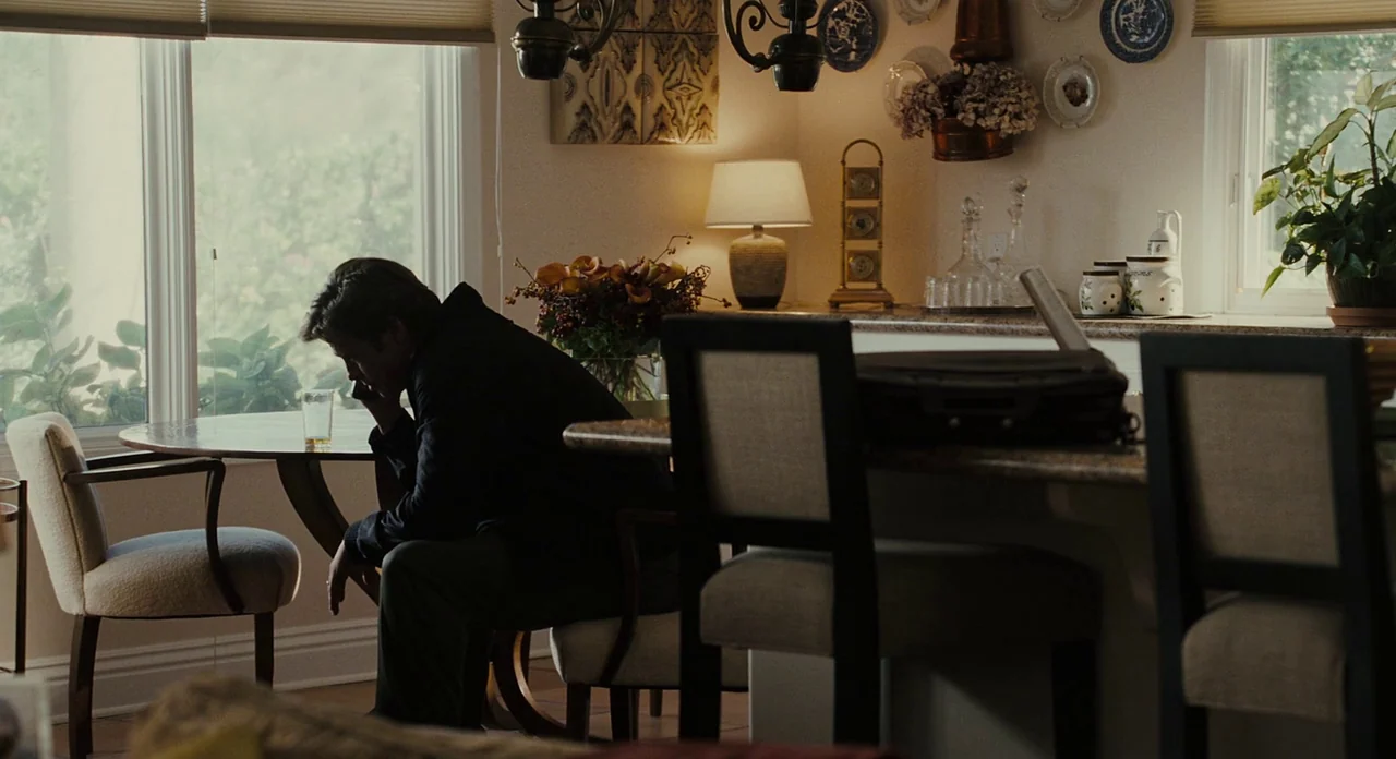 Moneyball (2011) movie still: medium — A residential dining area features a round wooden table, white chairs, and decorative wall plates. A man sits…