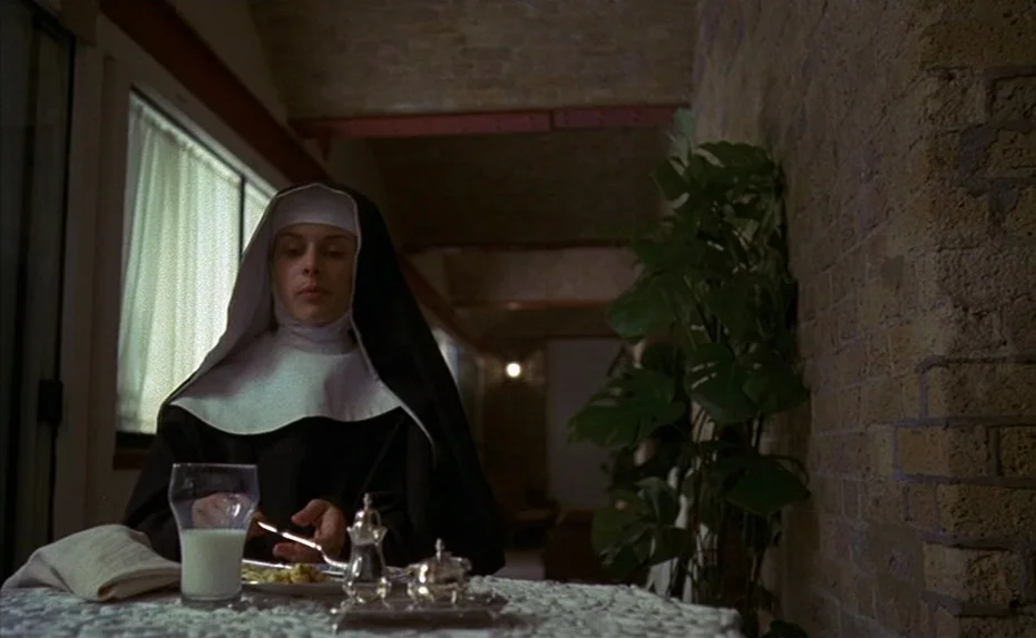 To The Devil a Daughter (1976) movie still: medium — A woman dressed as a nun sits at a table inside a room with brick walls. She holds a silver…
