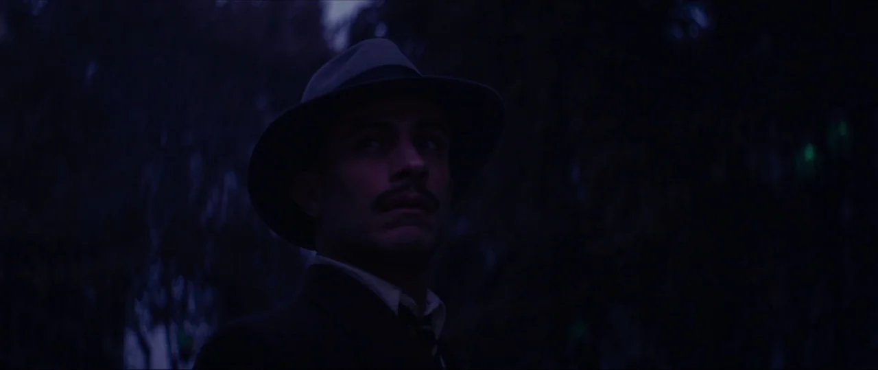 Neruda (2016) movie still: closeup — A nighttime scene in a dark, wooded park area obscured by heavy shadow. A man wearing a fedora looks upward…